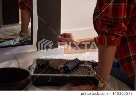 Woman applies dark paint along wall edge with small brush near tray and roller Woman applies dark paint along wall edge with small brush near tray and roller 140303572