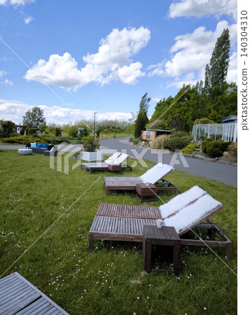 Wooden sun loungers, covered with cozy white throws, stand on the grass by the path. Wooden sun loungers, covered with cozy white throws, stand on the grass by the path. 140304310
