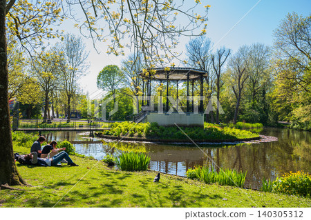 Historic park bandstand pavilion by pond with young people relaxing on green grass, beautiful sunny day in Vondelpark Amsterdam, spring landscape concept Historic park bandstand pavilion by pond with young people relaxing on green grass, beautiful sunny day in Vondelpark Amsterdam, spring landscape concept 140305312