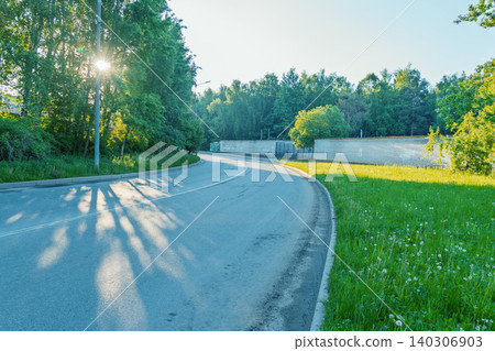 Sunlit curving road along a grassy verge and wooded edge Sunlit curving road along a grassy verge and wooded edge 140306903