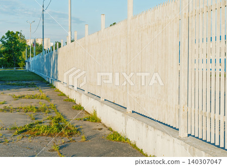 Long pale wooden slatted fence along cracked pavement with weeds 140307047
