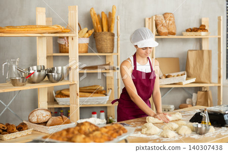 Teenager girl works as baker, mold baguette from dough, creates long loafs from dough. Teenager girl works as baker, mold baguette from dough, creates long loafs from dough. 140307438
