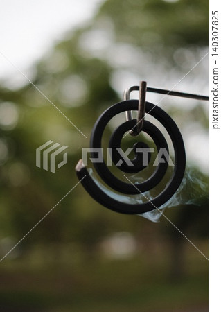 A hanging mosquito coil emits smoke against a backdrop of green trees. A hanging mosquito coil emits smoke against a backdrop of green trees. 140307825