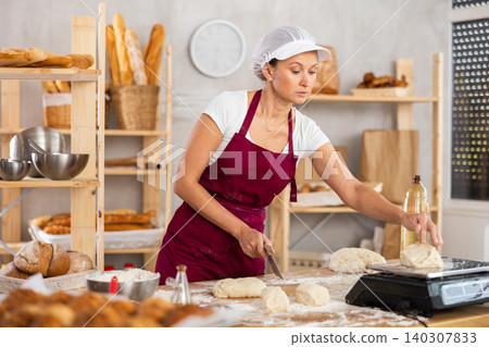 Middle-aged female baker cutting and weighing dough on scales in bakehouse Middle-aged female baker cutting and weighing dough on scales in bakehouse 140307833