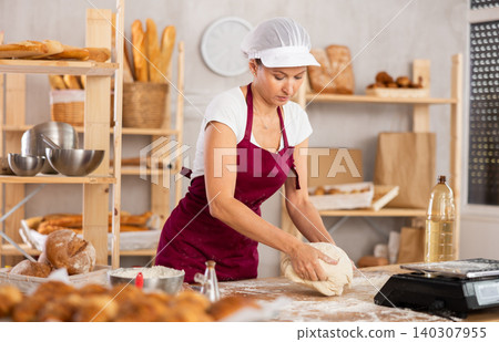 Young baker woman is standing near work table in kitchen, working with dough 140307955