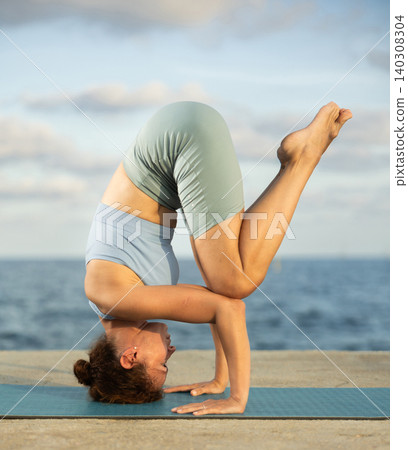 Sporty woman practitioner of yoga performing headstand, maintaining poised control and showcasing flexibility Sporty woman practitioner of yoga performing headstand, maintaining poised control and showcasing flexibility 140308304