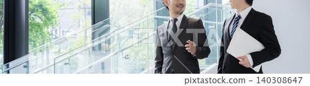 A young businessman and his boss converse by the window in a lush green office, communicating with a client (faceless banner). A young businessman and his boss converse by the window in a lush green office, communicating with a client (faceless banner). 140308647