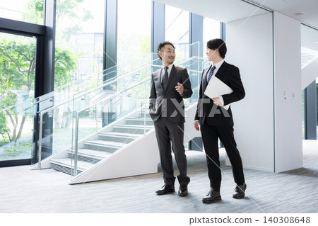 A young businessman and his superior converse by the window in the office surrounded by fresh greenery; the scene depicts communication with clients, business negotiations, and on-the-job training (OJT). A young businessman and his superior converse by the window in the office surrounded by fresh greenery; the scene depicts communication with clients, business negotiations, and on-the-job training (OJT). 140308648