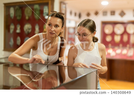 Young woman with teen daughter visitors stands near horizontal glass display case 140308649