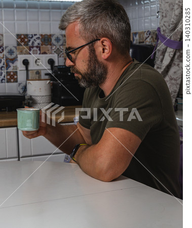 Man enjoys a cup of coffee while sitting at his kitchen table Man enjoys a cup of coffee while sitting at his kitchen table 140310285