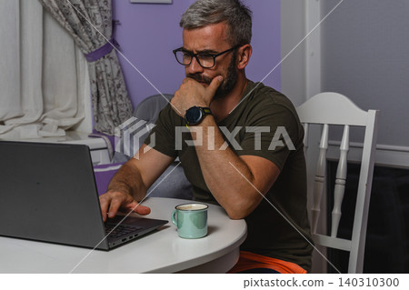 Man enjoys a cup of coffee while working on a laptop at home Man enjoys a cup of coffee while working on a laptop at home 140310300