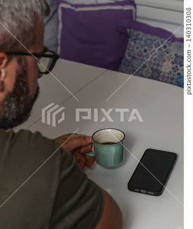 Man enjoys coffee while sitting at a table in a cozy home setting Man enjoys coffee while sitting at a table in a cozy home setting 140310308