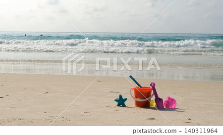 Children's colorful beach sand toy set including star mold, shovel, bucket and rake placed on sandy shore with sea waves behind. Play concept. Copy space Children's colorful beach sand toy set including star mold, shovel, bucket and rake placed on sandy shore with sea waves behind. Play concept. Copy space 140311964