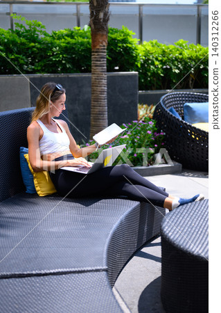 Woman working on laptop computer and reading notebook outdoor lounge, relaxed remote work lifestyle Woman working on laptop computer and reading notebook outdoor lounge, relaxed remote work lifestyle 140312266