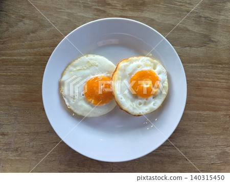 High angle view of two sunny-side-up fried eggs on a white round plate placed on a wooden table. High angle view of two sunny-side-up fried eggs on a white round plate placed on a wooden table. 140315450