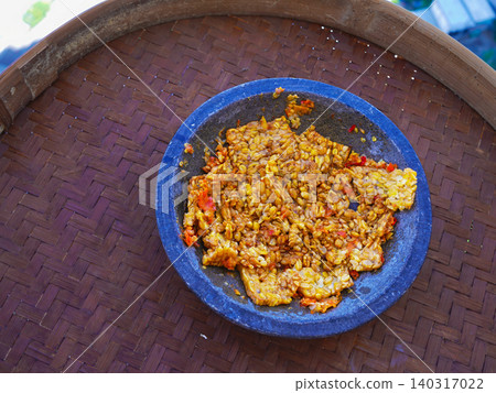 Traditional Indonesian food Tempe Penyet, smashed fried soybean cake with spicy chili sambal served in a rustic stone mortar on a woven bamboo tray. 140317022