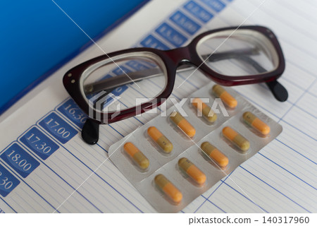 Close-up of reading glasses and a set of pills on a page of a paper daily planner, symbolizing treatment regimen for elderly people. Close-up of reading glasses and a set of pills on a page of a paper daily planner, symbolizing treatment regimen for elderly people. 140317960