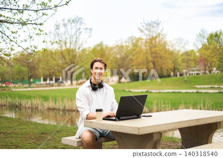 Smiling man enjoying remote freelance work on his laptop at an outdoor park table with coffee and headphones. Smiling man enjoying remote freelance work on his laptop at an outdoor park table with coffee and headphones. 140318540