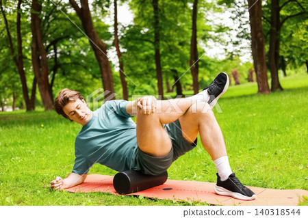 Man doing foam rolling for myofascial release and self-massage on a mat in a park to relieve muscle tension during post-workout recovery. Man doing foam rolling for myofascial release and self-massage on a mat in a park to relieve muscle tension during post-workout recovery. 140318544