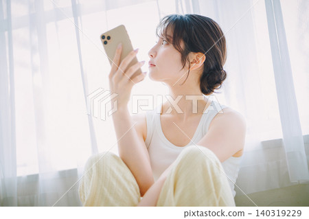 一位年輕女子在窗邊看著她的智慧型手機。 140319229