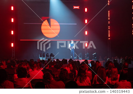Business presenter on stage under spotlight delivering keynote to seated audience at red lit conference Business presenter on stage under spotlight delivering keynote to seated audience at red lit conference 140319443