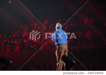 Speaker on stage addressing large audience in dimly lit auditorium during live event and performance Speaker on stage addressing large audience in dimly lit auditorium during live event and performance 140319445