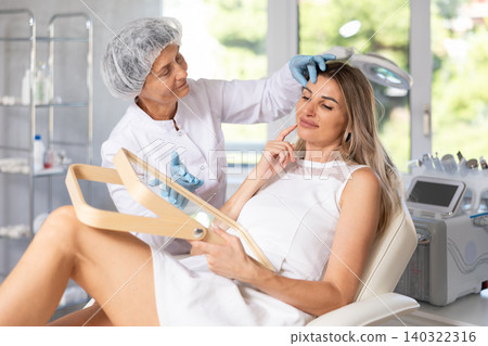 Young woman looking in mirror while mature cosmetologist showing results of facial procedure Young woman looking in mirror while mature cosmetologist showing results of facial procedure 140322316