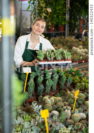 In store, positive gardening female worker inspects young cactus plants after treatment 140322433