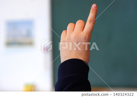 Child hand raised in classroom in front of chalkboard in the school classroom. Student rising index finger, indicating a question or participation Child hand raised in classroom in front of chalkboard in the school classroom. Student rising index finger, indicating a question or participation 140324322