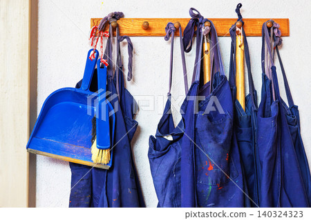 A row of blue aprons and a plastic dustpan with a small broom hanging on a wooden wall mounted hook inside a classroom or art room. A row of blue aprons and a plastic dustpan with a small broom hanging on a wooden wall mounted hook inside a classroom or art room. 140324323