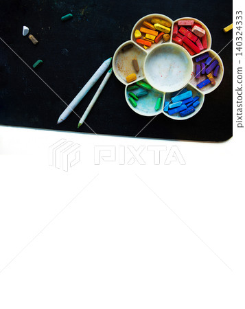 Top view of colorful pastel chalks arranged in a flower shaped palette with blending tools, placed on a black background with a white lower section Top view of colorful pastel chalks arranged in a flower shaped palette with blending tools, placed on a black background with a white lower section 140324333