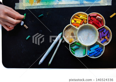 Top view of a hand arranging colorful pastel chalks in rainbow palette with blending tools on a black table Top view of a hand arranging colorful pastel chalks in rainbow palette with blending tools on a black table 140324336