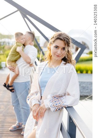 A Woman Dressed in a Flowing White Gown Stands Elegantly on a Picturesque Bridge, With Her Loving Husband Gently Holding Their Son by Her Side A Woman Dressed in a Flowing White Gown Stands Elegantly on a Picturesque Bridge, With Her Loving Husband Gently Holding Their Son by Her Side 140325308