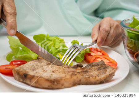 Asian elderly woman patient eating pork chop stake and vegetable salad for healthy food in hospital. Asian elderly woman patient eating pork chop stake and vegetable salad for healthy food in hospital. 140325994