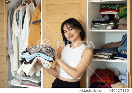Happy young Asian girl holding some items of clothing by the wardrobe Happy young Asian girl holding some items of clothing by the wardrobe 140327110