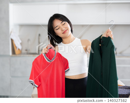 Happy young Asian girl trying to choose a dress in the kitchen 140327382