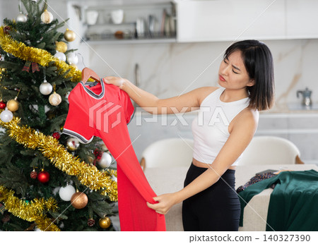 Sad young Asian girl trying to choose a dress standing next to Xmas tree Sad young Asian girl trying to choose a dress standing next to Xmas tree 140327390