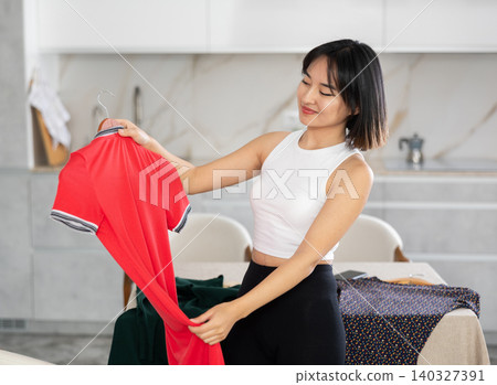 Happy young Asian girl trying to choose a dress in the kitchen Happy young Asian girl trying to choose a dress in the kitchen 140327391