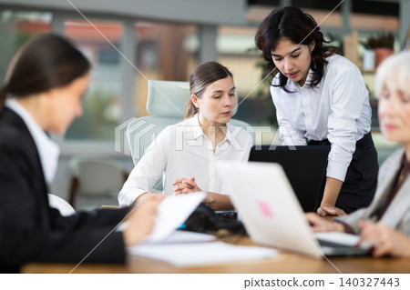 Asian girl leaned over to seated woman employee, helps with work and looks at laptop screen Asian girl leaned over to seated woman employee, helps with work and looks at laptop screen 140327443