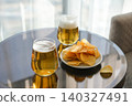 Two glasses of cold light beer with foam next to a plate of ruffled potato chips on reflective glass table. Minimalist lifestyle shot of appetizers and drinks in bright setting 140327491