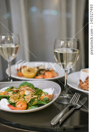 Elegant dinner for two featuring shrimp salads with almond flakes, lemon and glasses of white wine on dark reflective table in hotel room. Concept of luxury dining and romantic evening Elegant dinner for two featuring shrimp salads with almond flakes, lemon and glasses of white wine on dark reflective table in hotel room. Concept of luxury dining and romantic evening 140327493