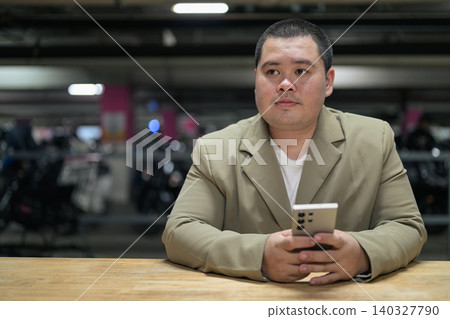 Pensive plus size Isan Thai businessman holding phone in parking garage Pensive plus size Isan Thai businessman holding phone in parking garage 140327790