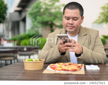 Focused plus size Isan Thai businessman using phone with pizza outdoors Focused plus size Isan Thai businessman using phone with pizza outdoors 140327821
