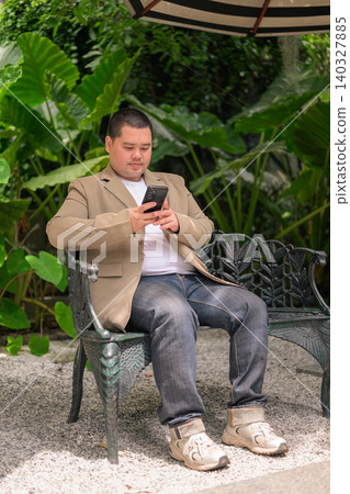 Plus size Isan Thai businessman using phone on garden bench vertical 140327885
