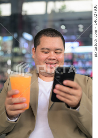 Smiling plus size Isan Thai businessman using phone with orange juice vertical Smiling plus size Isan Thai businessman using phone with orange juice vertical 140327936
