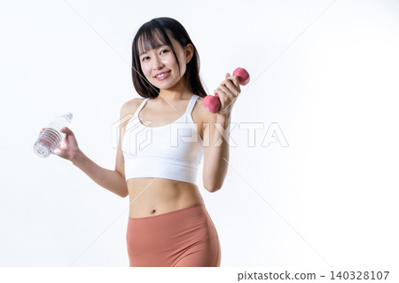 A woman with a smile working out at the gym, holding water and dumbbells. (Image of a woman training) A woman with a smile working out at the gym, holding water and dumbbells. (Image of a woman training) 140328107