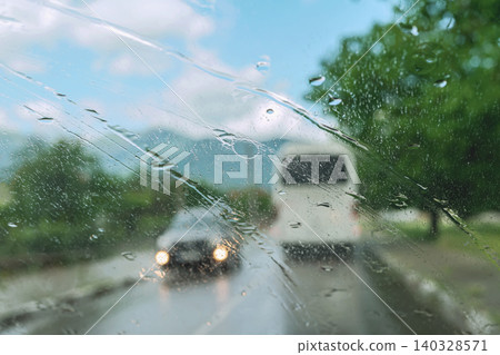 Rain drops cover car windshield with blurred traffic, road, trees, sky, and vehicle headlights beyond glass. Natural background with windshield water for weather, transport, travel, driving themes 140328571