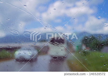 Wet car windshield covered with rain droplets shows blurred houses, road, sky, and clouds beyond glass. Natural background with windshield moisture for weather, travel, transport, driving themes Wet car windshield covered with rain droplets shows blurred houses, road, sky, and clouds beyond glass. Natural background with windshield moisture for weather, travel, transport, driving themes 140328572