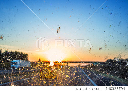 Blurry highway through a smudged dirty car windshield, affecting visibility during sunset, emphasizing highway traffic safety concerns and the importance of clean glass for safe travel. Blurry highway through a smudged dirty car windshield, affecting visibility during sunset, emphasizing highway traffic safety concerns and the importance of clean glass for safe travel. 140328794