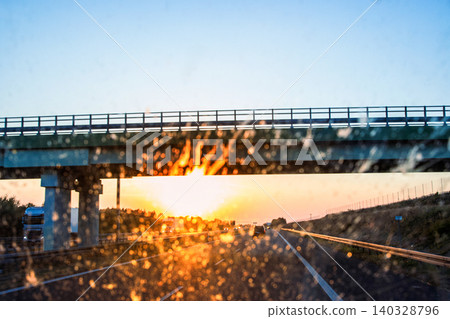 Highway and bridge at sunset through a dirty windshield, emphasizing road safety and the challenges of visibility while driving on long journeys. Transportation, logistics, road traffic. Copy space Highway and bridge at sunset through a dirty windshield, emphasizing road safety and the challenges of visibility while driving on long journeys. Transportation, logistics, road traffic. Copy space 140328796